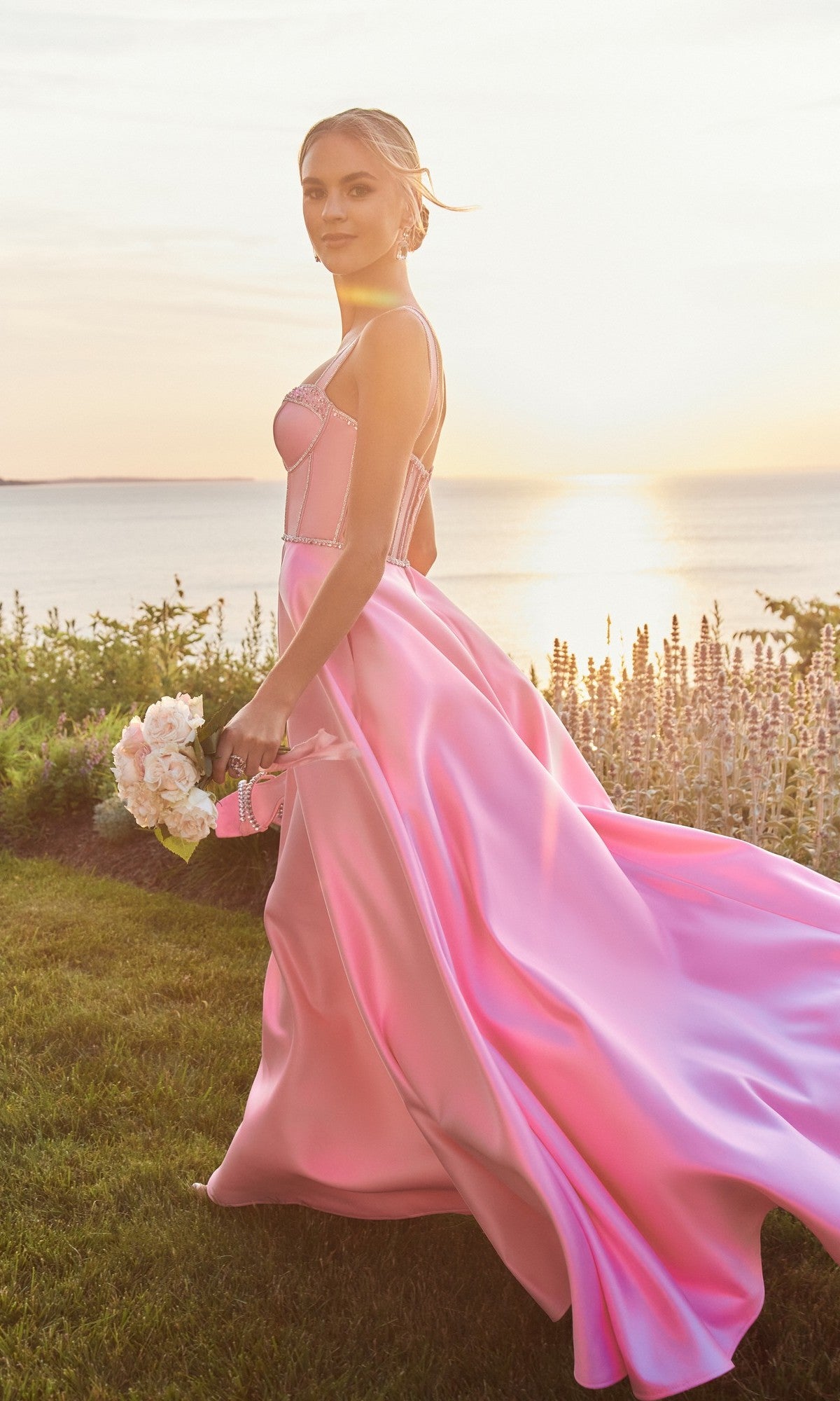 A woman wearing the Morilee 49177 long prom dress with a beaded corset bodice holds a bouquet as she walks outdoors near a body of water at sunset.